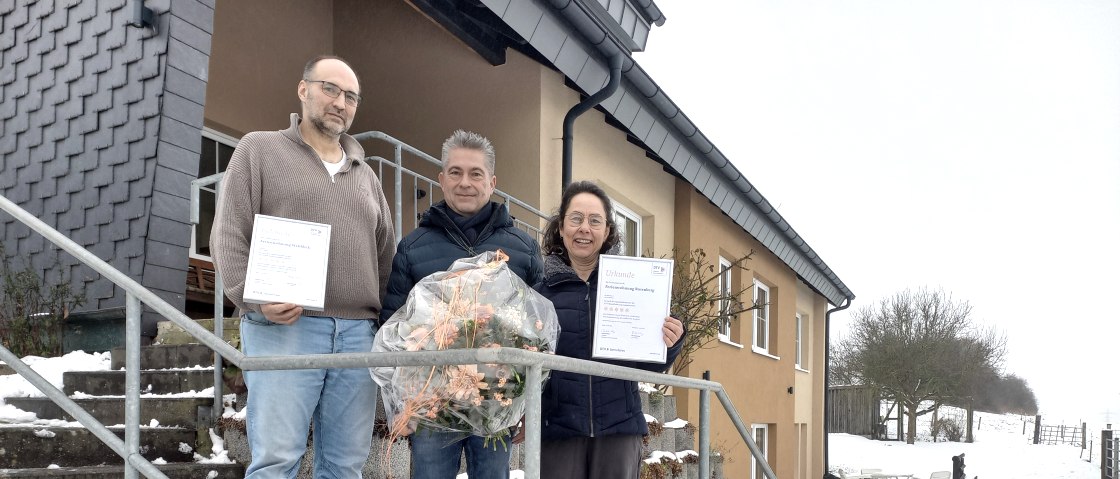 Das Bild zeigt drei Personen, die vor einem Haus im Schnee stehen. Die beiden &auml;u&szlig;eren Personen halten Urkunden in der Hand, w&auml;hrend die mittlere Person einen Blumenstrau&szlig; tr&auml;gt. Vor ihnen steht ein Schild mit der Aufschrift "Rosenberghof Vulkaneifel Ferienwohnung" und einem QR-Code. 