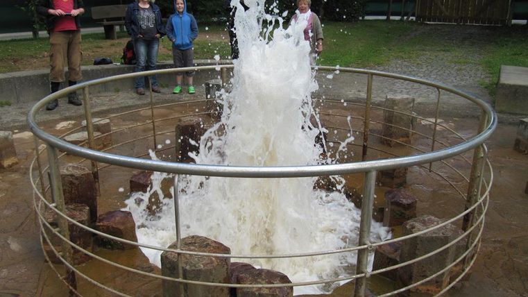 Ein Wasserspielplatz mit spritzendem Wasser in einem runden Becken. Im Hintergrund stehen mehrere Personen, die das Schauspiel beobachten.