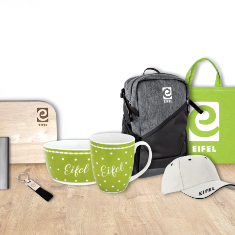 Ein Brettchen, Flachmann, Schl&uuml;sselanh&auml;nger, Schale, Tasse, Rucksack, Stoffbeutel und Basecap nebeneinander auf einem Holzboden. Alle Produkte sind aus dem Eifelshop und mit dem Schriftzug Eifel und/oder dem Logo "e" versehen.