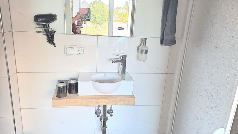 Ein modernes Badezimmer mit einem Waschbecken auf einem Holzregal und einem großen Spiegel. Im Hintergrund ist eine Dusche und ein Fenster zu sehen.
