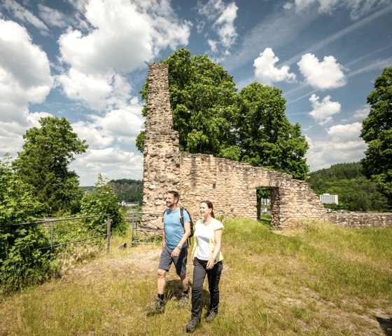 Zwei Wanderer spazieren an einer alten Steinmauer vorbei, umgeben von gr&uuml;nen B&auml;umen und einem blauen Himmel mit Wolken., &copy; Eifel Tourismus GmbH, Dominik Ketz