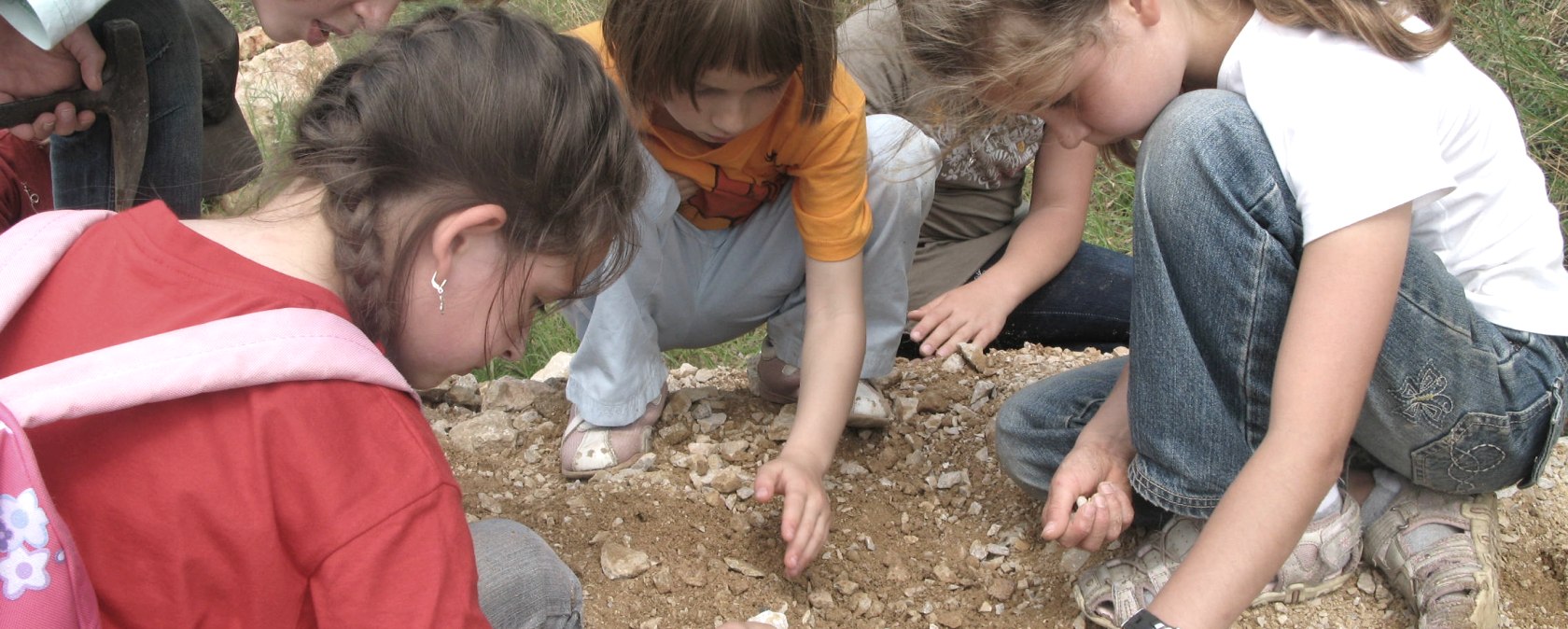 6 children kneel in a circle on a mound of rocks and look at them closely.
