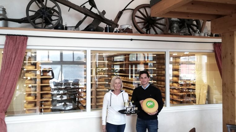 Ein Mann und eine Frau stehen vor eine großen Glasscheibe, hinter der in Regalen zahlreiche Käselaibe reifen. Dei Frau trägt ein Tablett mit Getränken und der Mann einen Käselaib.