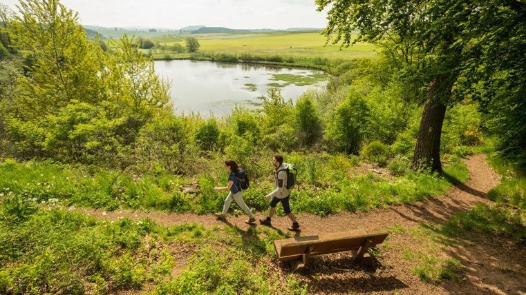 Twee wandelaars op een pad bij Eichholzmaar in de Eifel, omgeven door groene natuur en een meer op de achtergrond.