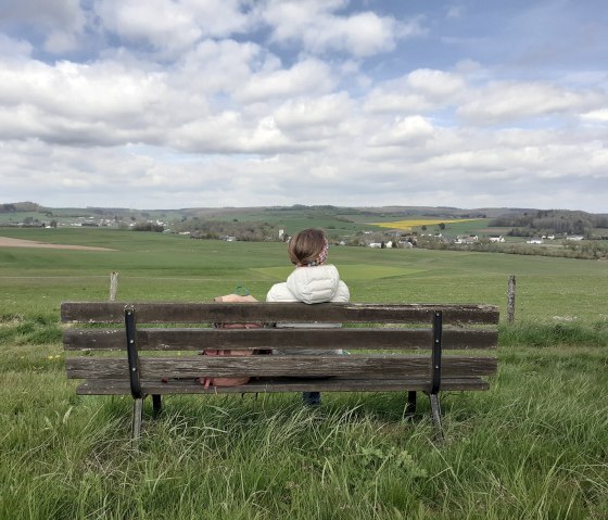 A person sits on a bench and looks out over a wide, green landscape with fields and clouds in the sky., © Touristik GmbH Gerolsteiner Land, Ute Klinkhammer