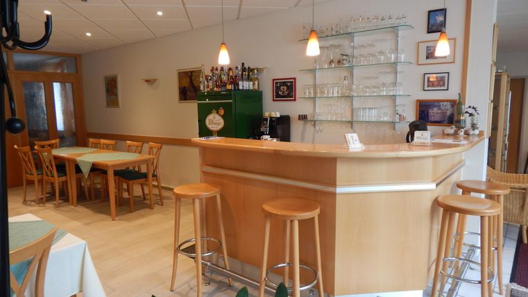 Eine einladende Bar mit Holztheke und modernen Barhockern. Der Raum ist hell erleuchtet und hat eine gemütliche Atmosphäre.