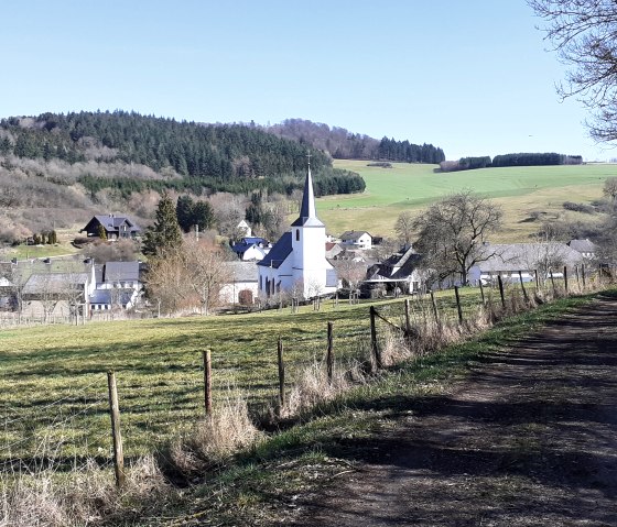 Village view of Rockeskyll with church in the foreground, surrounded by green fields and forests under a blue sky., © Touristik GmbH Gerolsteiner Land, Ute Klinkhammer