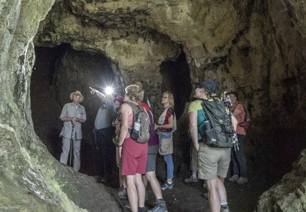 A hiking group of around 10 people stands in the interior of a large cave. The people are marvelling at the massive stone walls around them.