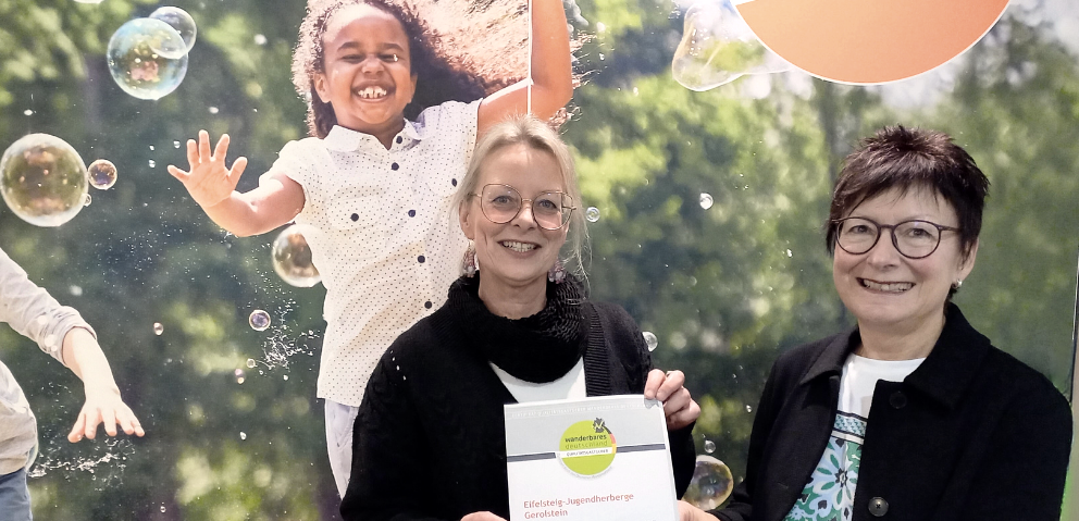 Das Foto zeigt zwei Frauen, die stolz ein Zertifikat halten, vor einem Hintergrund, der eine junge, fr&ouml;hliche Person in Seifenblasen darstellt.Die Szene zeigt zwei Frauen, die sich f&uuml;r ein Foto aufgestellt haben, wobei eine das Zertifikat h&auml;lt, das sie pr&auml;sentieren. Der Hintergrund zeigt eine gro&szlig;formatige Darstellung eines Kindes, das inmitten von Seifenblasen fr&ouml;hlich in die Kamera l&auml;chelt. Der Hintergrund ist ein gr&uuml;ner, natur&auml;hnlicher Hintergrund. Die Frauen stehen vor dem Hintergrund, wobei die Frau rechts das Zertifikat f&uuml;r die Kamera h&auml;lt, w&auml;hrend die Frau links ebenfalls beteiligt ist. Die Ausrichtung des Fotos ist gut und das Hauptaugenmerk liegt auf den Frauen und dem Zertifikat. Der Hintergrund dient als erg&auml;nzende Kulisse.Die beiden Hauptfiguren sind zwei Frauen mittleren Alters. Die Frau links hat blondes Haar, tr&auml;gt eine Brille und einen schwarzen Pullover. Sie h&auml;lt das Zertifikat mit einem L&auml;cheln. Die Frau rechts hat kurz geschnittenes, braunes Haar, eine Brille und einen dunklen Blazer. Sie l&auml;chelt ebenfalls. Der Hintergrund zeigt ein gl&uuml;ckliches Kind, das in Seifenblasen spielt., &copy; Touristik GmbH Gerolsteiner Land