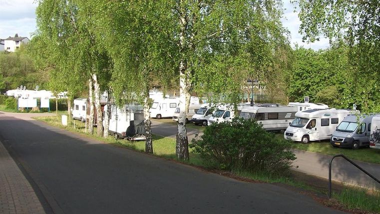 Wohnmobilstellplatz in Gerolstein mit mehreren Wohnmobilen unter grünen Bäumen. Asphaltierte Straße und Gehweg im Vordergrund.