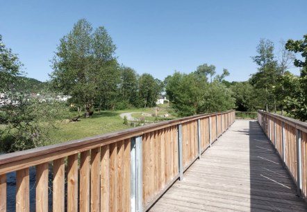 Eine gro&szlig;e Holzbr&uuml;cke f&uuml;hrt in ein Parkgel&auml;nde mit zahlreichen B&auml;umen und Spazierwegen.