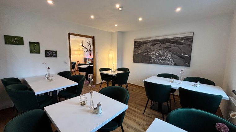 Ein modernes Restaurant mit hellen Wänden und eleganten, runden Tischen. Die Stühle sind grün und eine große Wandkunst ziert den Raum.