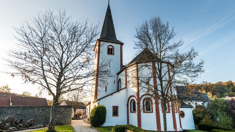 Kirche mit Kirchturm  in weiß gehalten mit ziegelsteinroten Akzenten, im Vordergrund Wiese mit zwei Bäumen, gutes Wetter