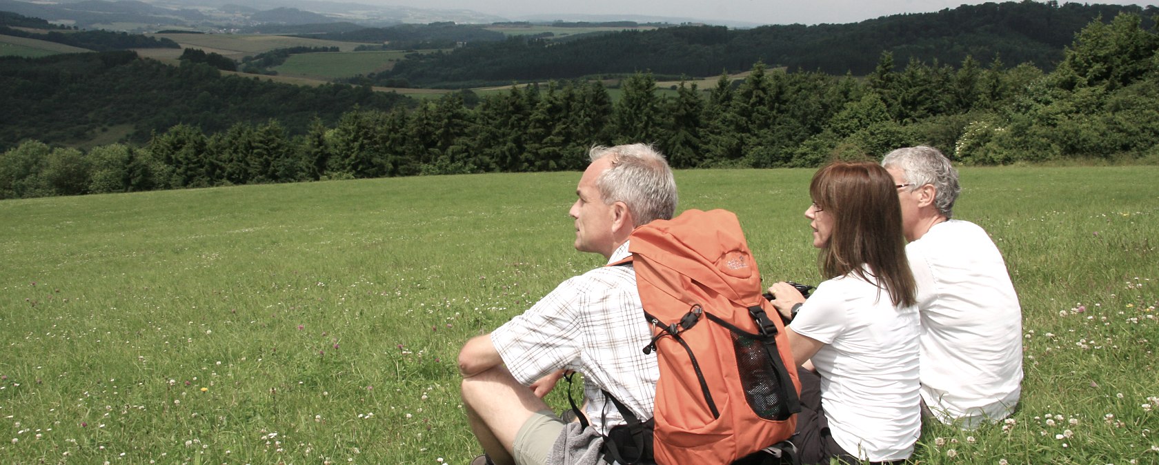 Drie wandelaars zitten op een groene weide en staren in de verte. Een van hen draagt een oranjekleurige rugzak. Op de achtergrond zijn heuvels en bossen te zien., © Eifel Tourismus GmbH