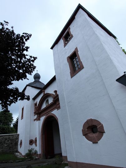 Schloss Oberehe mit weißer Fassade, roten Fensterrahmen und einem Turm. Der Himmel ist bewölkt, ein Baum ist im Vordergrund sichtbar.