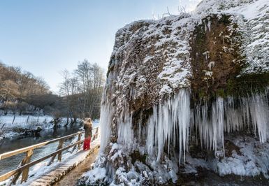 Gefrorener Nohner Wasserfall mit Eiszapfen, eine Person auf einem Steg, winterliche Landschaft mit Schnee und kahlen Bäumen
