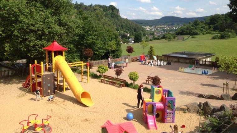 Ein bunter Spielplatz mit Rutschen, Schaukeln und Spielhäusern. Im Hintergrund sind Bäume und eine grüne Landschaft zu sehen.