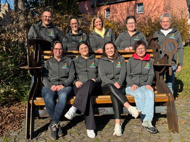 Eine Gruppe aus 9 Personen, die z.T. auf einer Bank sitzen, schauen lachend in die Kamera. Alle Personen tragen die gleiche Jacke mit eingesticktem Firmenlogo.