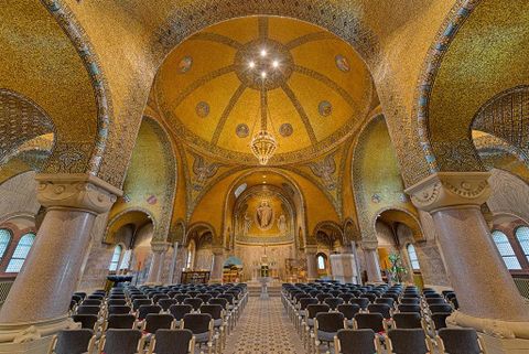 Un intérieur majestueux avec des mosaïques dorées et de hautes arches.
Des rangées de chaises sont disposées, et un autel est visible à l'arrière-plan.