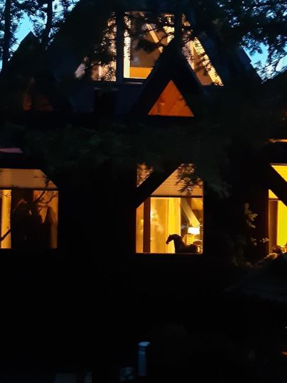 Ein gemütliches Haus mit einem spitzen Dach und beleuchteten Fenstern. Umgeben von Bäumen in der Dämmerung.