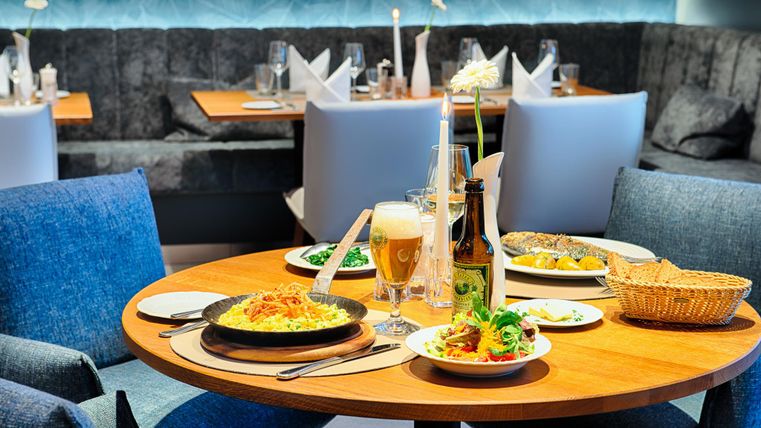 Ein Tisch in einem Restaurant mit verschiedenen Speisen, darunter Pasta, Salat und Fisch. Ein Bierglas und eine Flasche stehen daneben.