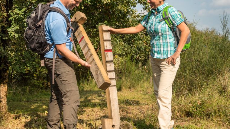 Zwei Wanderer betrachten ein hölzernes Kerbholz auf einem Wanderweg.