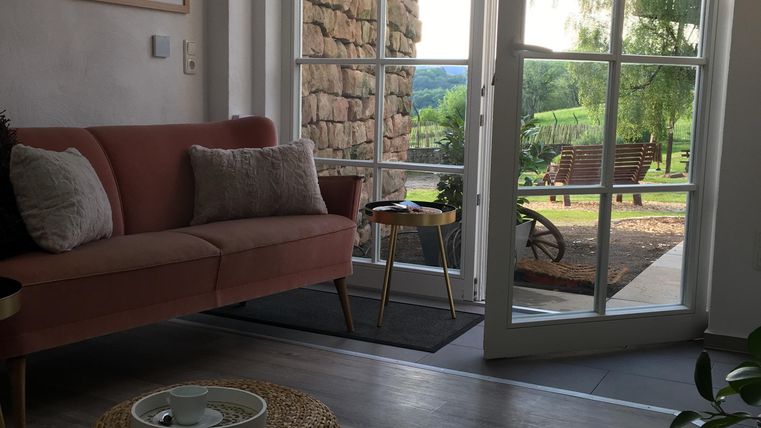 Un salon confortable avec un canapé rose et une vue sur la terrasse. La lumière entre par les grandes fenêtres et à l'extérieur, un paysage verdoyant est visible.
