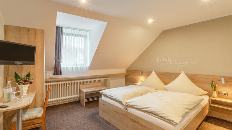 Ein gemütliches Hotelzimmer mit einem Doppelbett und einem kleinen Tisch. Die Wände sind hell und es gibt viel Tageslicht durch das Fenster.