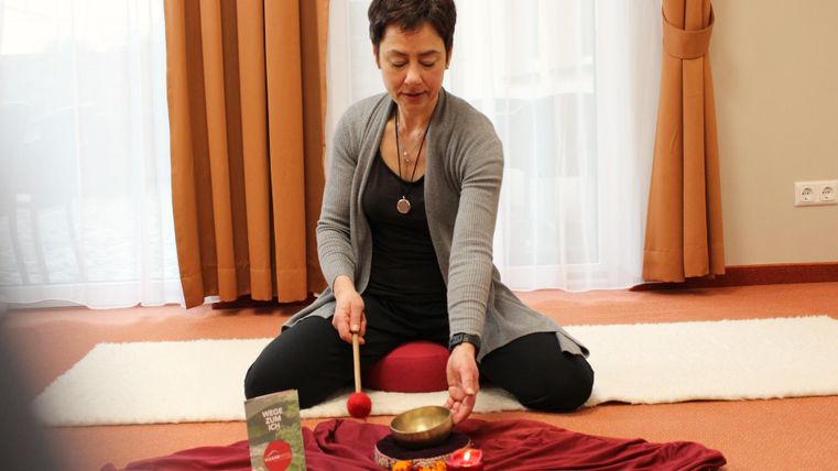 Une femme est assise en tailleur sur le sol et joue avec un bol chantant. Devant elle se trouve un tissu rouge avec différents objets.