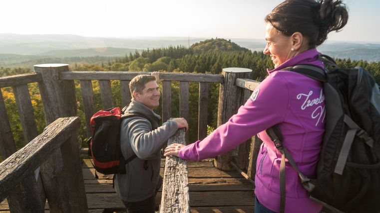 Zwei Personen auf einem Aussichtsturm mit Blick auf bewaldete Hügel im Hintergrund.