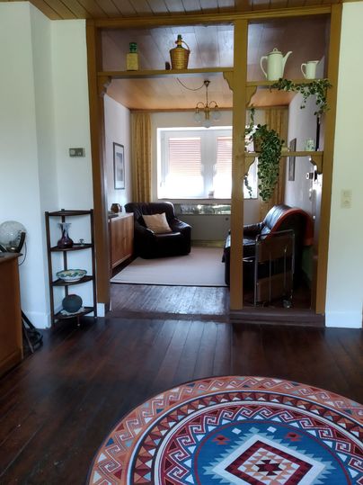 Une pièce cosy avec un tapis rond et deux fauteuils confortables. Par une ouverture, on peut voir un espace de vie lumineux.