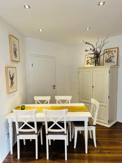 Een lichte eetruimte met een witte tafel en stoelen. Aan de muren hangen schilderijen, en er staat een elegante kast in de hoek.