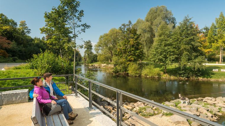 Ein Paar sitzt auf einer Bank im Kurpark, blickt auf einen Fluss. Ein Hund steht am Ufer. Umgeben von Bäumen und blauem Himmel.