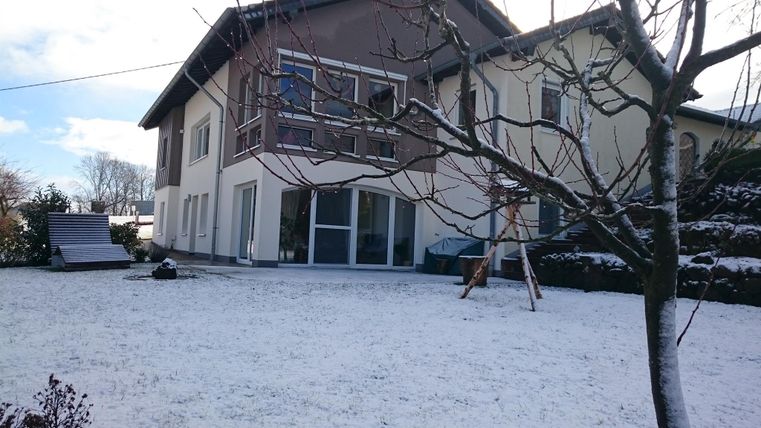 Ein gemütliches Haus im Schnee, umgeben von einem gepflegten Garten. Der Himmel ist teilweise bewölkt, und die Landschaft wirkt ruhig und winterlich.