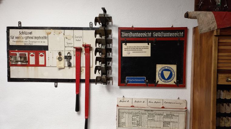 Ein Schlüsselbrett mit Werkzeugen und eine Unterrichtstafel hängen an einer weißen Wand. Daneben sind Schilder und ein Holzschrank zu sehen.