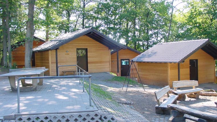 Drei Holzhütten im Wald, umgeben von Bäumen. Eine Tischtennisplatte und Holzbänke stehen auf einem gepflasterten Platz.