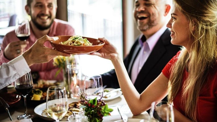 Eine Gruppe von Menschen sitzt in einem Restaurant, lacht und genießt gemeinsam ein Essen. Auf dem Tisch stehen Weingläser und Teller mit Speisen.