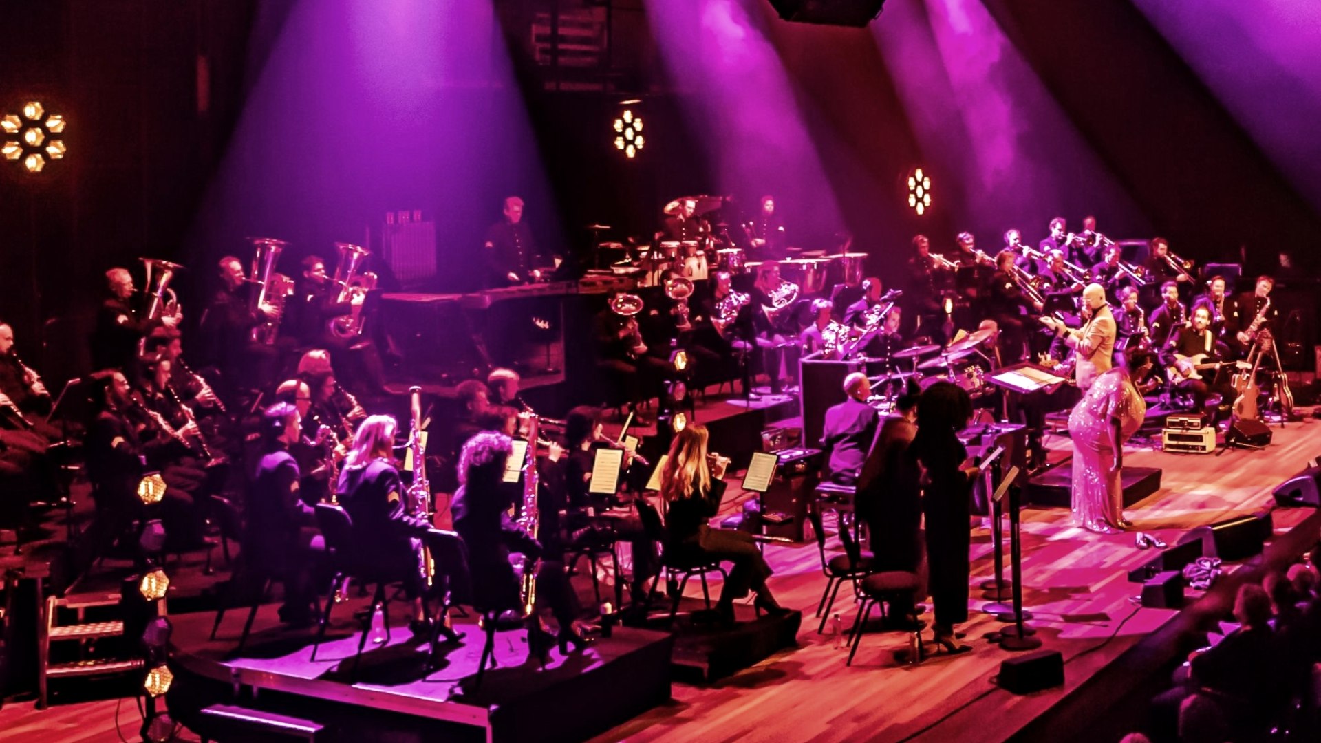 Das Bild zeigt ein Orchester bei einer Auff&uuml;hrung auf einer B&uuml;hne, die in einem lebhaften Magenta-Ton beleuchtet ist. Das Orchester besteht aus zahlreichen Musikern, die verschiedene Instrumente spielen, darunter Blasinstrumente, Streichinstrumente und Schlagzeug. Ein Dirigent steht vor dem Orchester, und eine S&auml;ngerin in einem glitzernden Kleid steht ebenfalls auf der B&uuml;hne. Im Vordergrund ist ein Teil des Publikums zu sehen, das der Auff&uuml;hrung zusieht. Die Beleuchtung tr&auml;gt zur dramatischen Atmosph&auml;re bei., &copy; Touristik GmbH Gerolsteiner Land