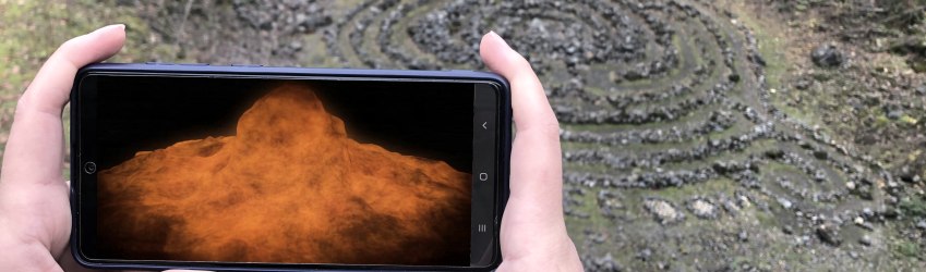 Two hands hold a smartphone across the abyss of a volcano. The volcanic explosion is visualised on the screen.