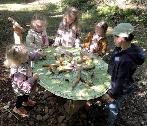 Fünf Kinder spielen an einem runden Tisch im Wald. Der Tisch hat ein grünes, baumartiges Muster mit Holzstücken darauf., © Touristik GmbH Gerolsteiner Land