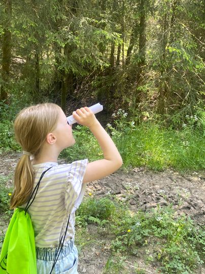 Ein Mädchen schaut mit einem Fernglas in den Wald. Sie trägt einen grünen Rucksack und steht auf einem naturbelassenen Weg.