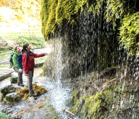 Zwei Personen stehen vor einem moosbedeckten Wasserfall. Sie tragen Outdoor-Kleidung und schauen fasziniert auf das herabfließende Wasser., © Eifel Tourismus GmbH, Dominik Ketz