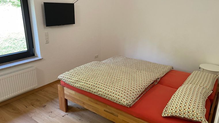 Une chambre simple avec un lit double et des coussins colorés. Un téléviseur est accroché au mur, et une fenêtre apporte de la lumière naturelle.