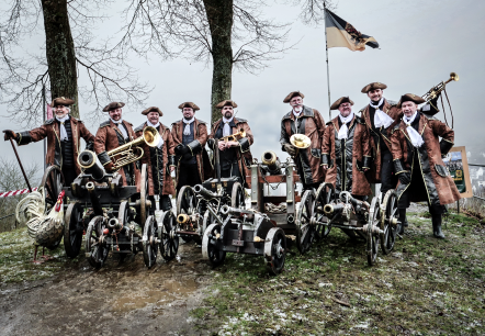 Op een foto poseert een groep mannen in historische kleding, omringd door kleine kanonnen en muziekinstrumenten, tegen de achtergrond van een somber winterlandschap.De sc&egrave;ne toont een groep van negen mannen die zich opstellen op een licht verhoogd grasveld, met voor zich meerdere kleine kanonnen en muziekinstrumenten zoals trompetten en een trombone. Ze lijken te poseren voor een groepsfoto of zich voor te bereiden op een optreden. De kanonnen staan in een rij opgesteld, met een decoratieve metalen haan aan de linkerkant. Op de achtergrond is een bewolkt landschap te zien met bomen en een heuvel met uitzicht op een stad, evenals een vlag met een wapen. De compositie is symmetrisch, waarbij de mannen en kanonnen een dominante horizontale lijn vormen, die in evenwicht wordt gehouden door de verticale elementen van de bomen en de vlag., &copy; Klaus Wollwert 