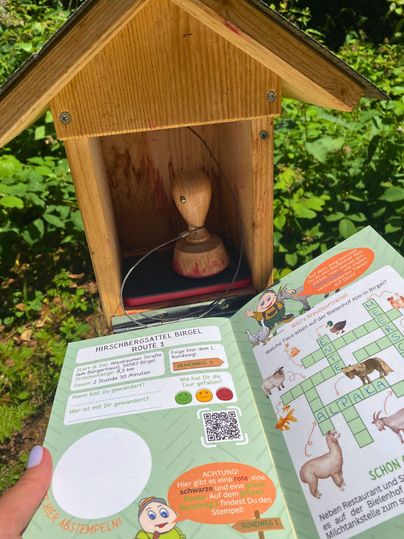 Ein Wanderpass mit Stempelstation im Freien, umgeben von grüner Vegetation.