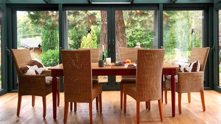 Een lichte serre met een houten tafel en vier stoelen. Het uitzicht toont een groene landschap door de grote ramen.