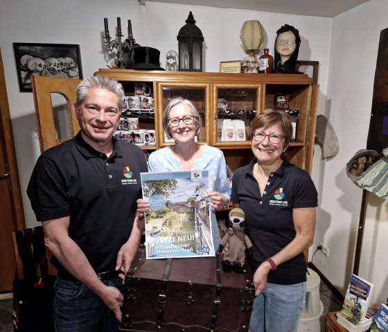 Drei lächelnde Personen stehen in einem kleinen Raum vor einem Holzschrank und halten eine große Schatzkiste und ein Plakat zum Detektiv-Trail hoch.