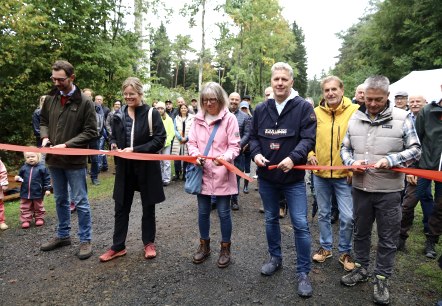 Five people with scissors in their hands cut a red ribbon at the same time. Behind them are numerous spectators and children.