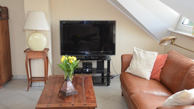Een gezellige woonkamer met een bruine bank en een houten tafel. Op de tafel staat een vaas met gele tulpen en op de achtergrond staat een televisie.