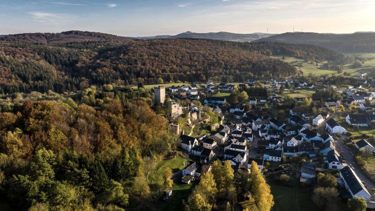 Luftaufnahme von Kerpen: Burg, umgeben von herbstlichen Wäldern und Häusern. Hügelige Landschaft im Hintergrund unter klarem Himmel.
